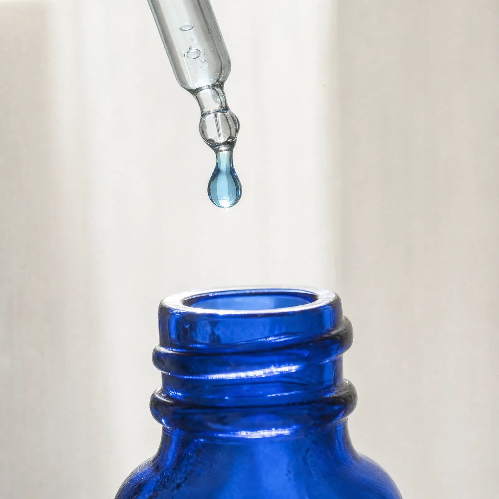 A glass pipette releasing a single blue drop above the open neck of a cobalt-blue glass bottle.