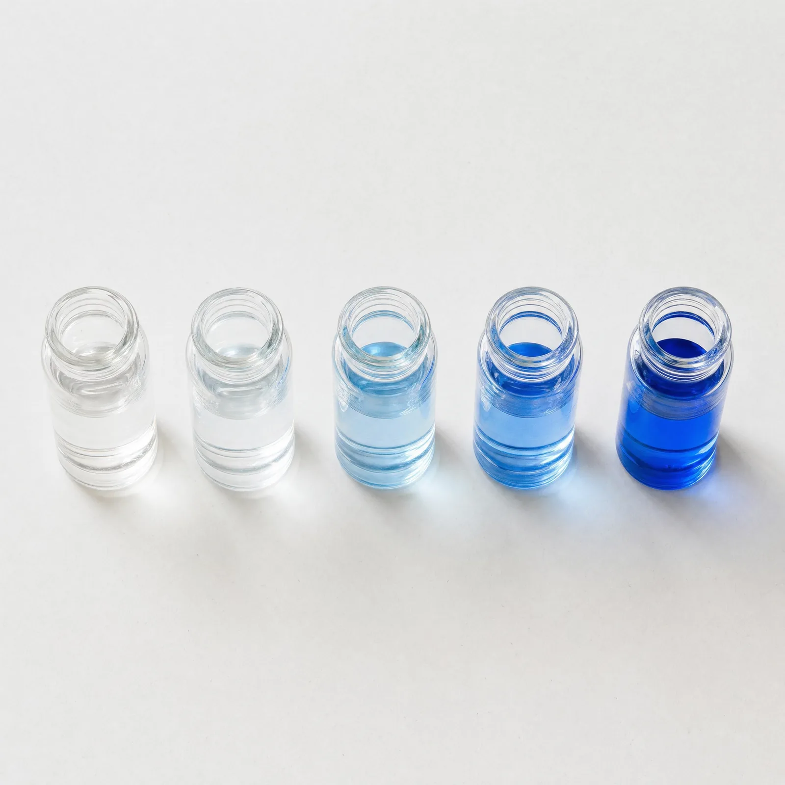 Five small glass vials in a row on a white surface, the liquid in them running from clear on the left to deep blue on the right.
